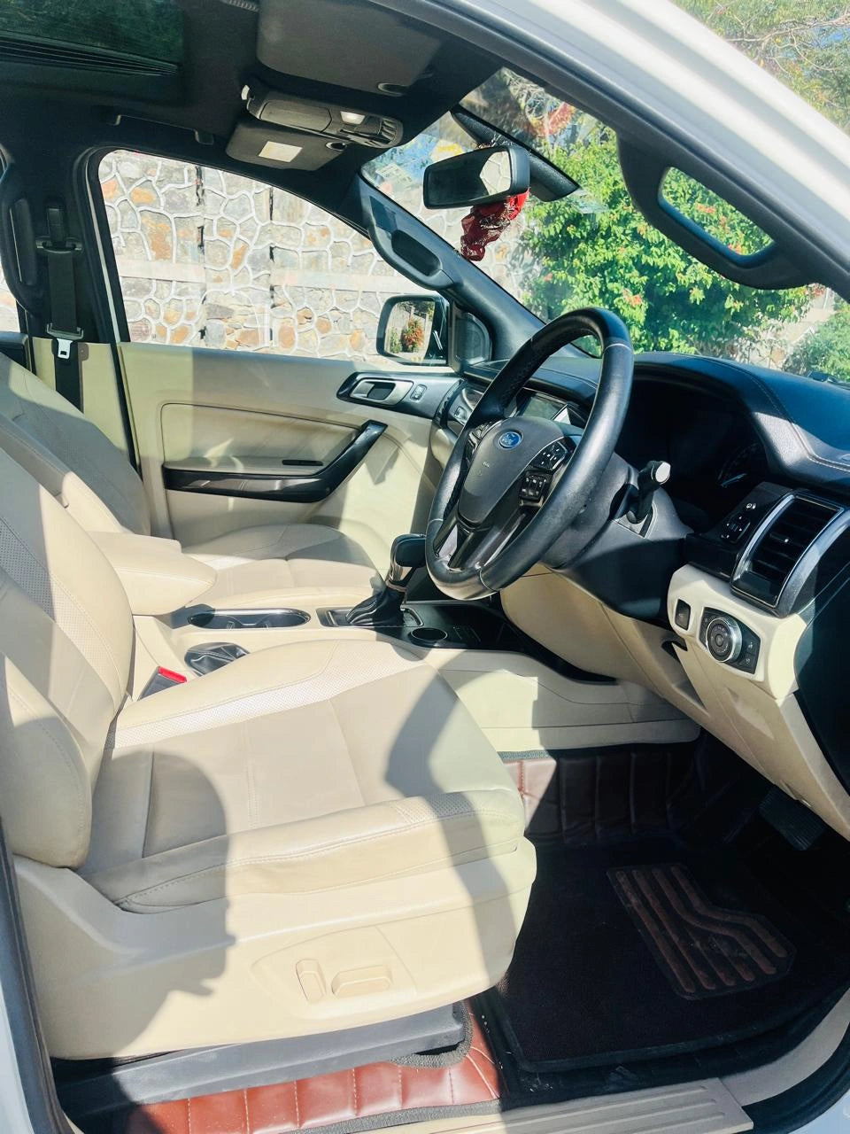 Car interior with beige seats and steering wheel, showing a stone wall and greenery outside.