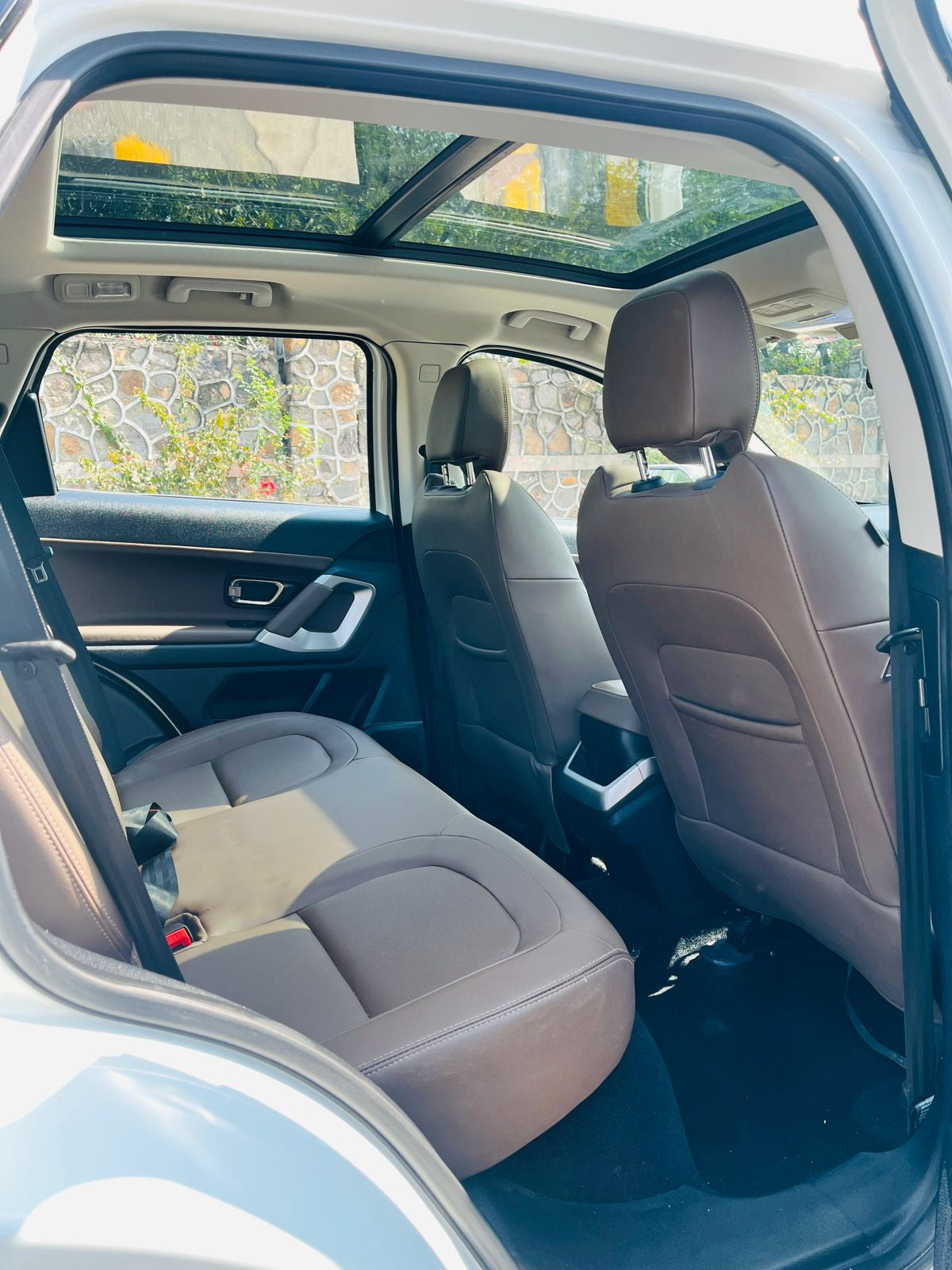 Backseat of a car with gray seats and a sunroof, showing a stone wall and greenery outside.