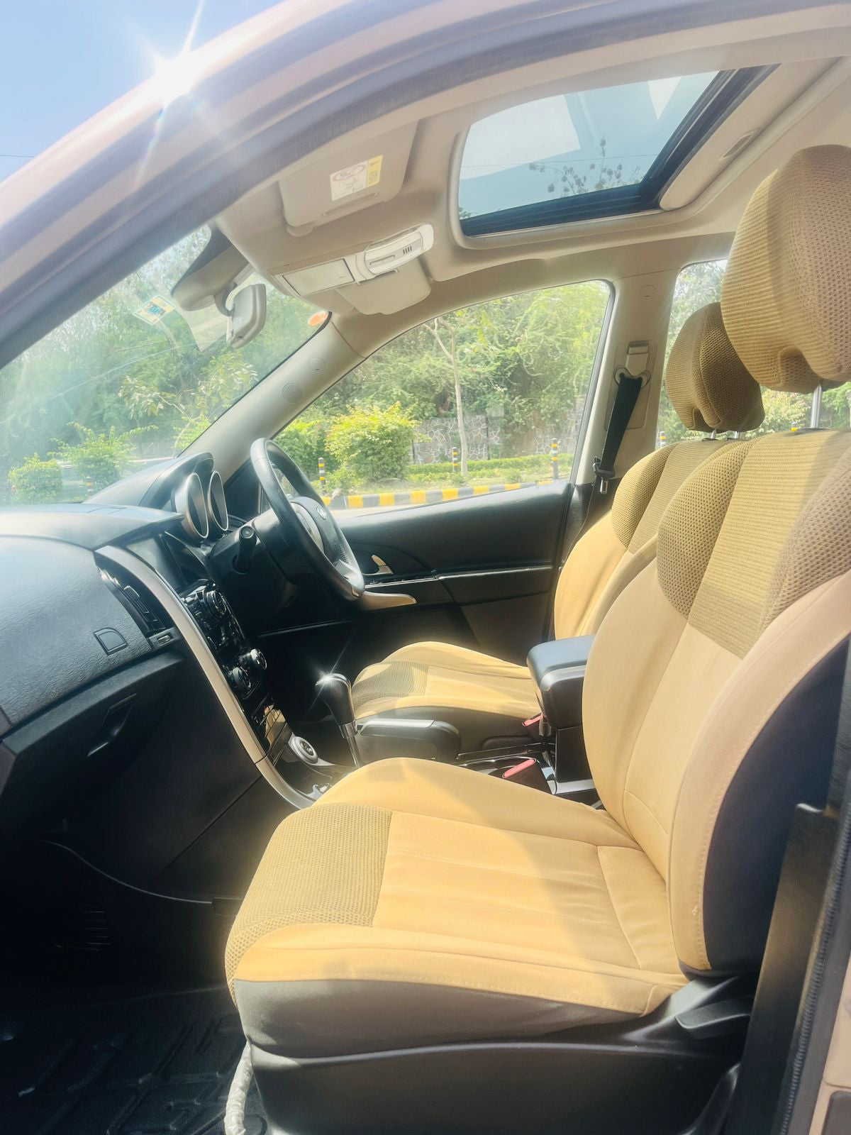 Car interior with beige seats and dashboard, sunroof open, showing greenery outside.